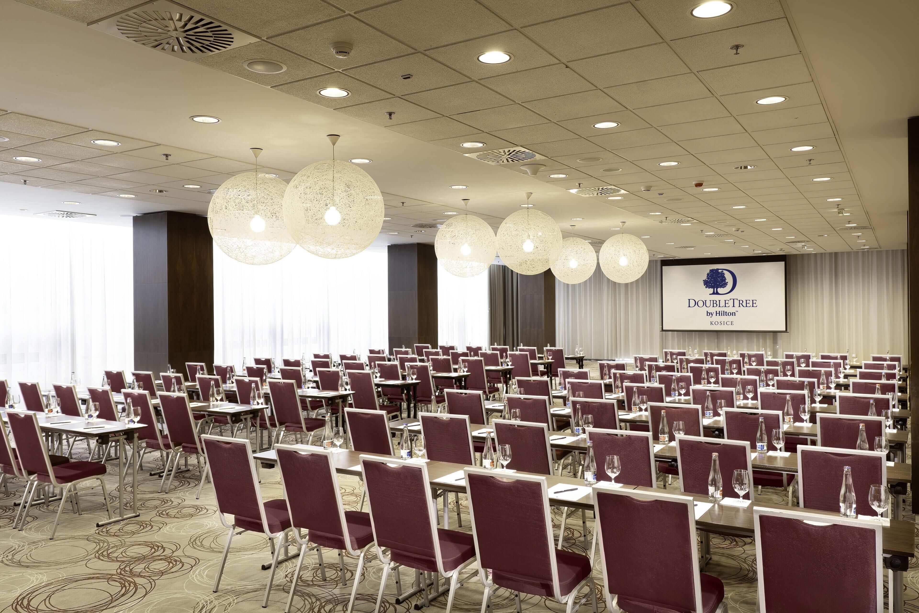 A conference room with rows of tables and chairs, large pendant lights, and a projection screen displaying the DoubleTree by Hilton logo.