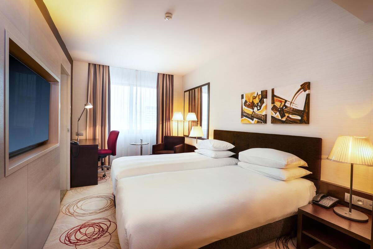 A hotel room with two beds, a desk, a chair, and artwork on the wall.