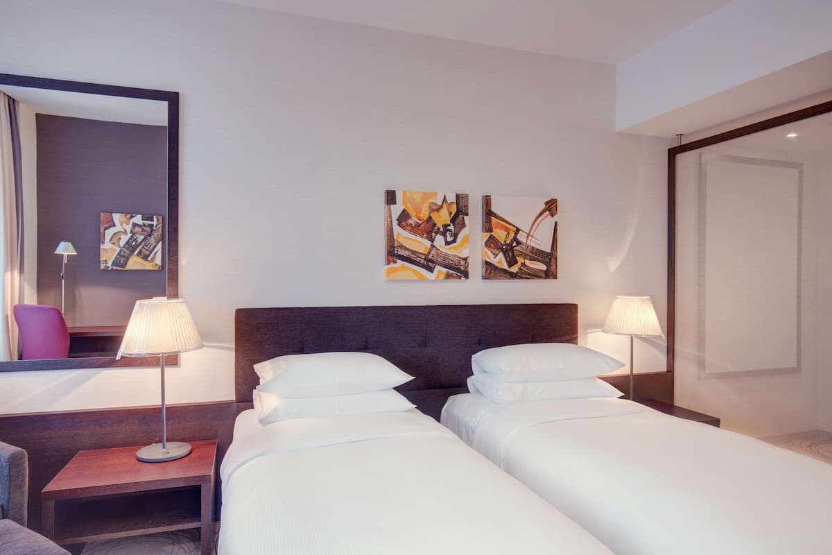 A hotel room with two beds, a nightstand with a lamp, a chair, and abstract artwork on the wall.