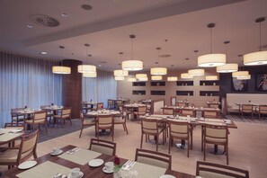 Uma área de refeições de hotel com várias mesas e cadeiras, iluminação pendente e uma grande janela com cortinas.