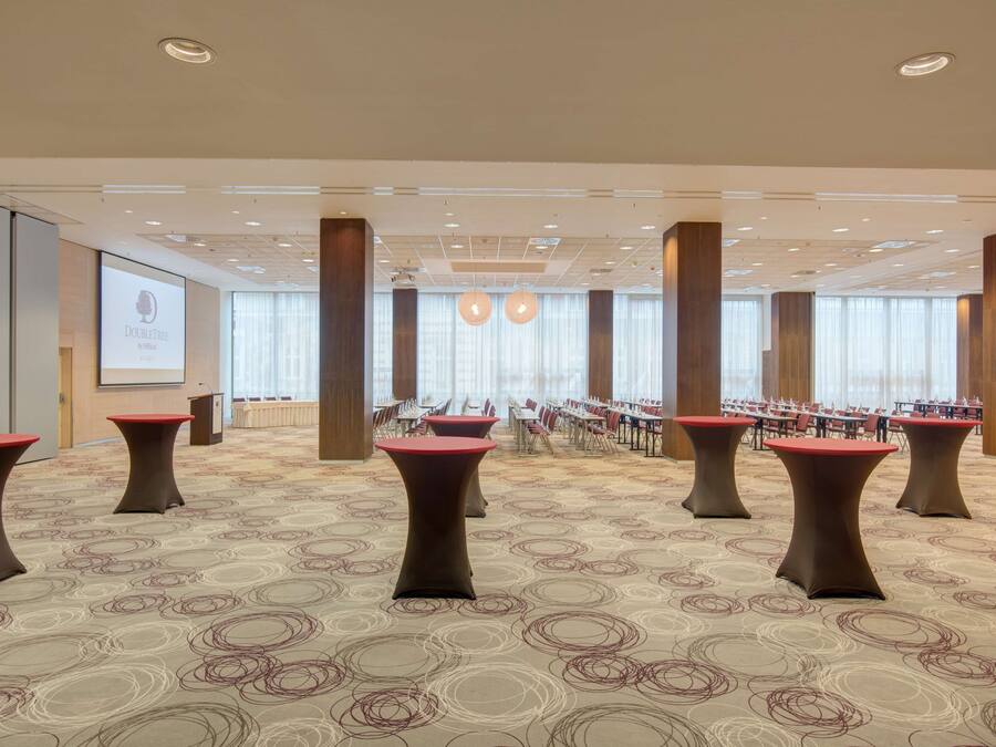Une salle de conférence équipée de plusieurs tables et chaises, d’un écran de projection et de grandes fenêtres avec des rideaux.