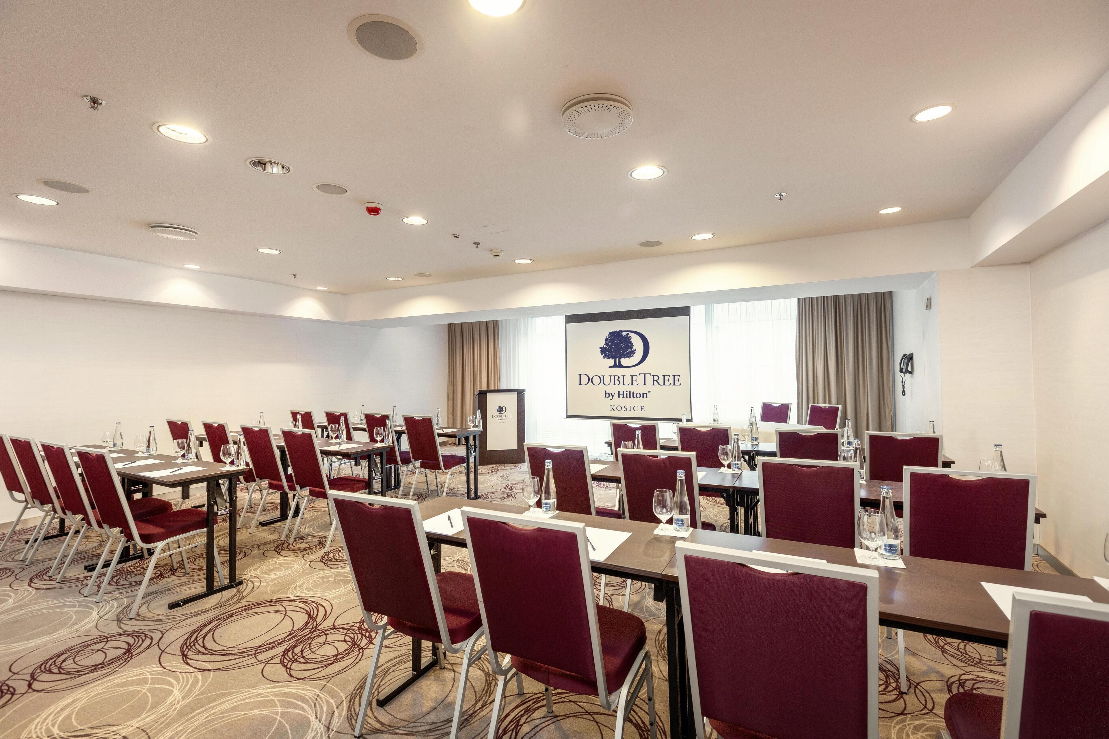 Uma sala de conferências com mesas, cadeiras, um projetor e um banner com a inscrição 'DOUBLETREE by Hilton Kosice'.
