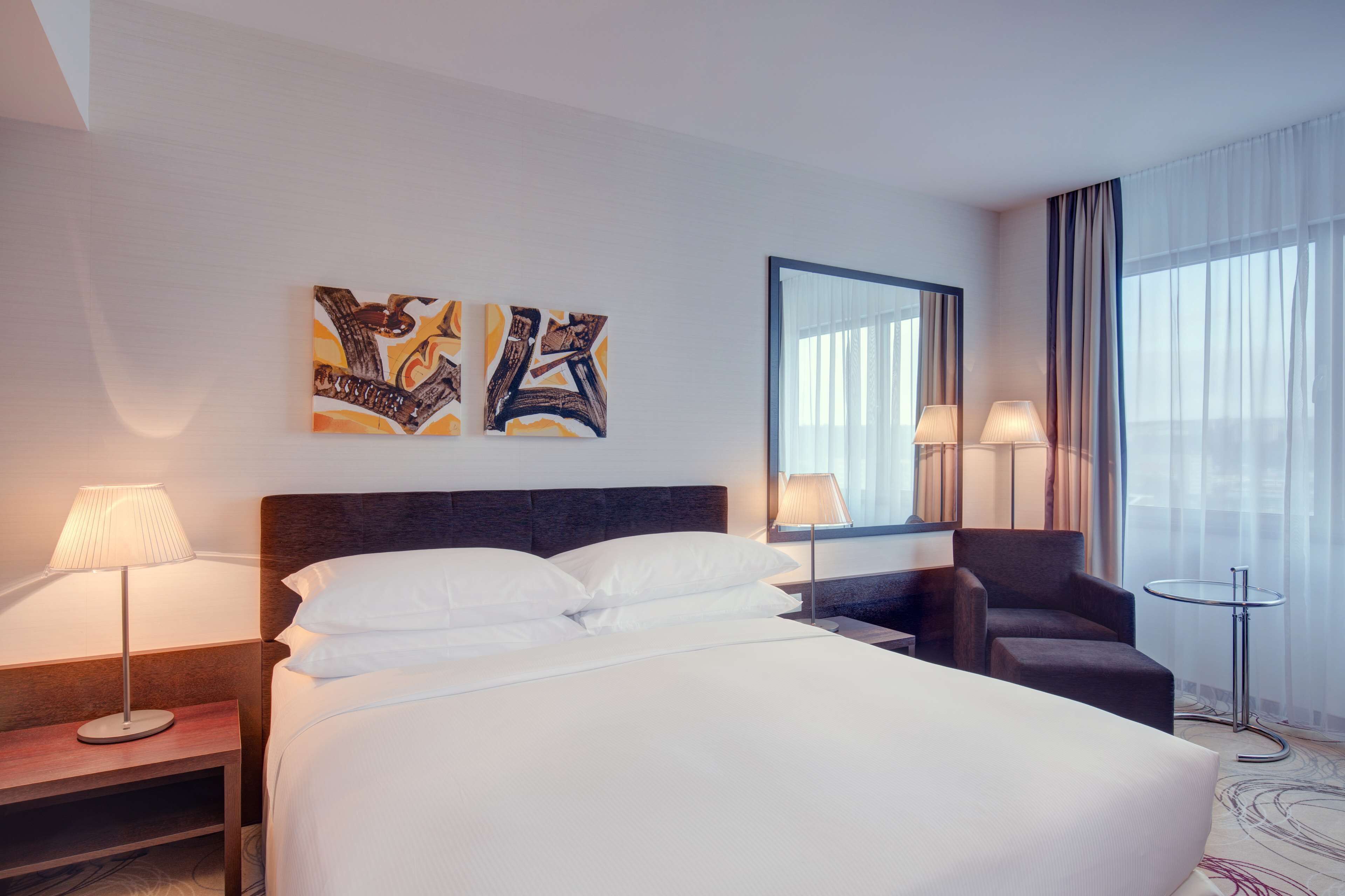 A hotel room with a large bed, two bedside tables with lamps, a chair, and a small table. There are abstract paintings on the wall and a large mirror above the bed.