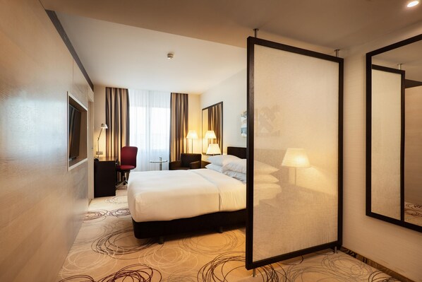Quarto de hotel com uma cama grande, uma escrivaninha com cadeira, televisão e um espelho amplo.