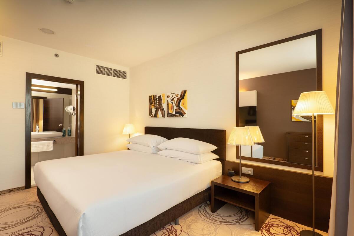 A hotel room with a large bed, a bedside table with a lamp, a mirror, and artwork on the wall.