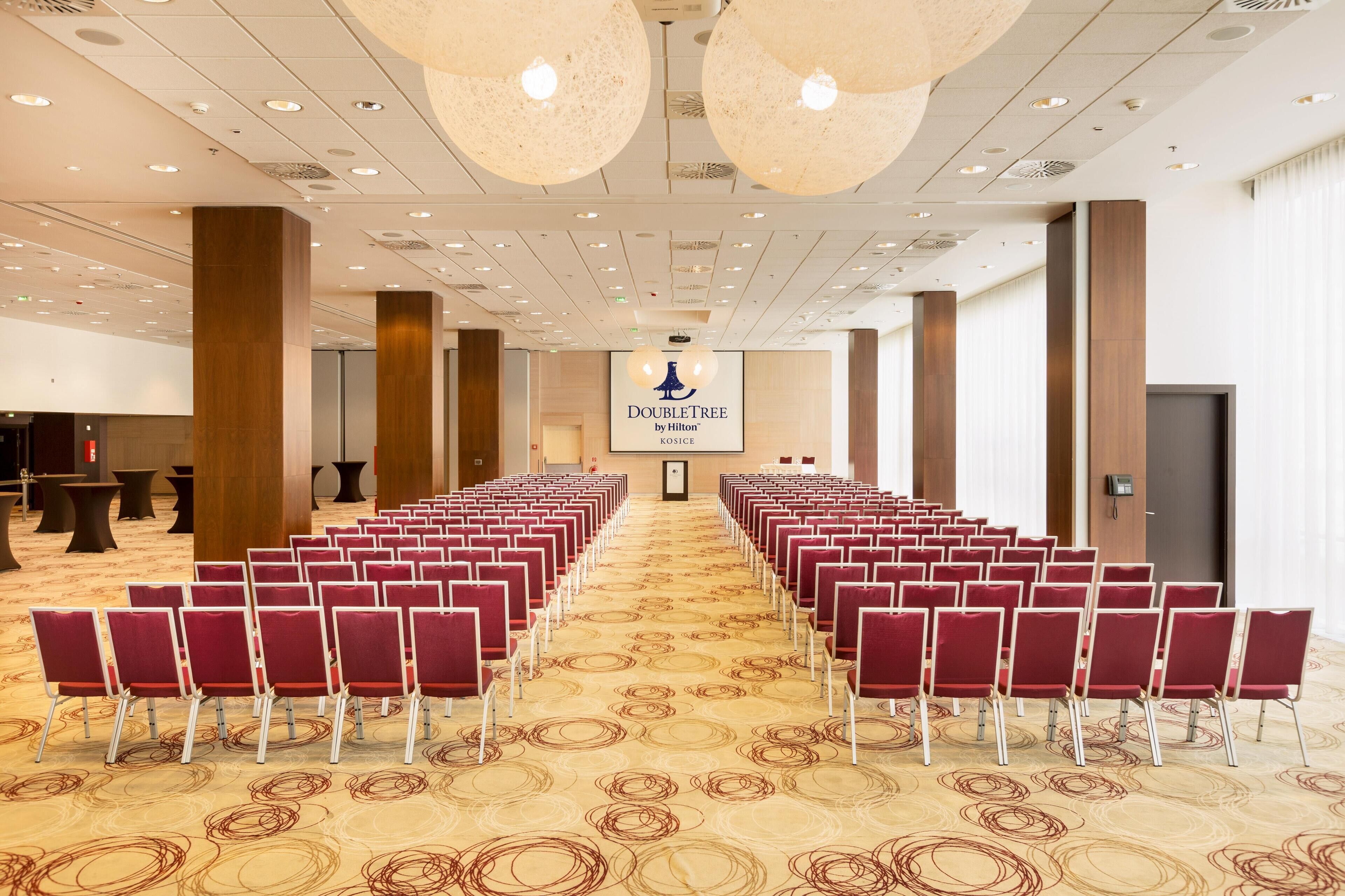 Un salón de conferencias con hileras de sillas rojas, un atril y una pantalla que muestra “DOUBLETREE by Hilton KOSICE”.