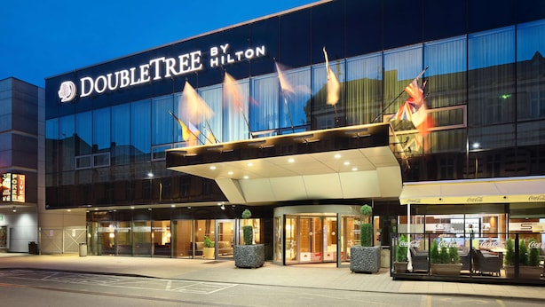 A DoubleTree by Hilton hotel with a modern glass facade, outdoor seating area, and a Coca-Cola branded entrance.
