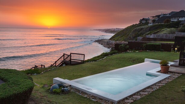 A swimming pool with a view of the ocean at sunset, a wooden deck, and a grassy area with a house in the background. - The Sands @ St Francis (Saint Francis Bay)