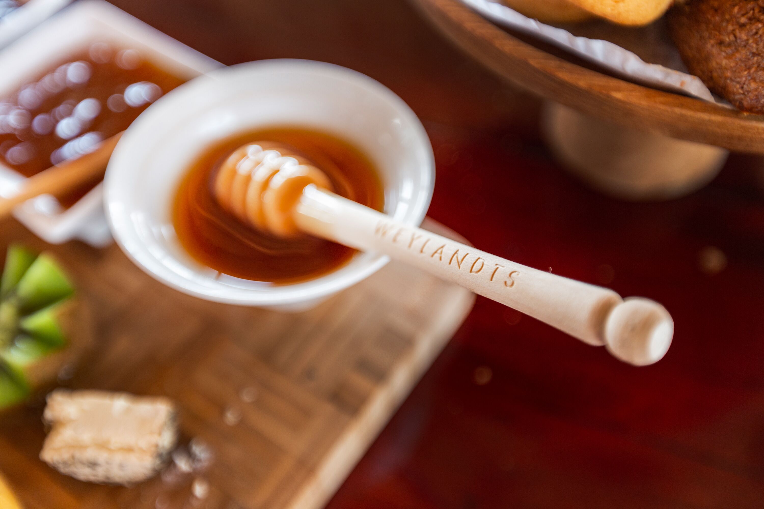 Un cucharón para miel con la inscripción "WEYLANDS", un pequeño cuenco de miel y una bandeja de madera con un muffin y un trozo de queso.