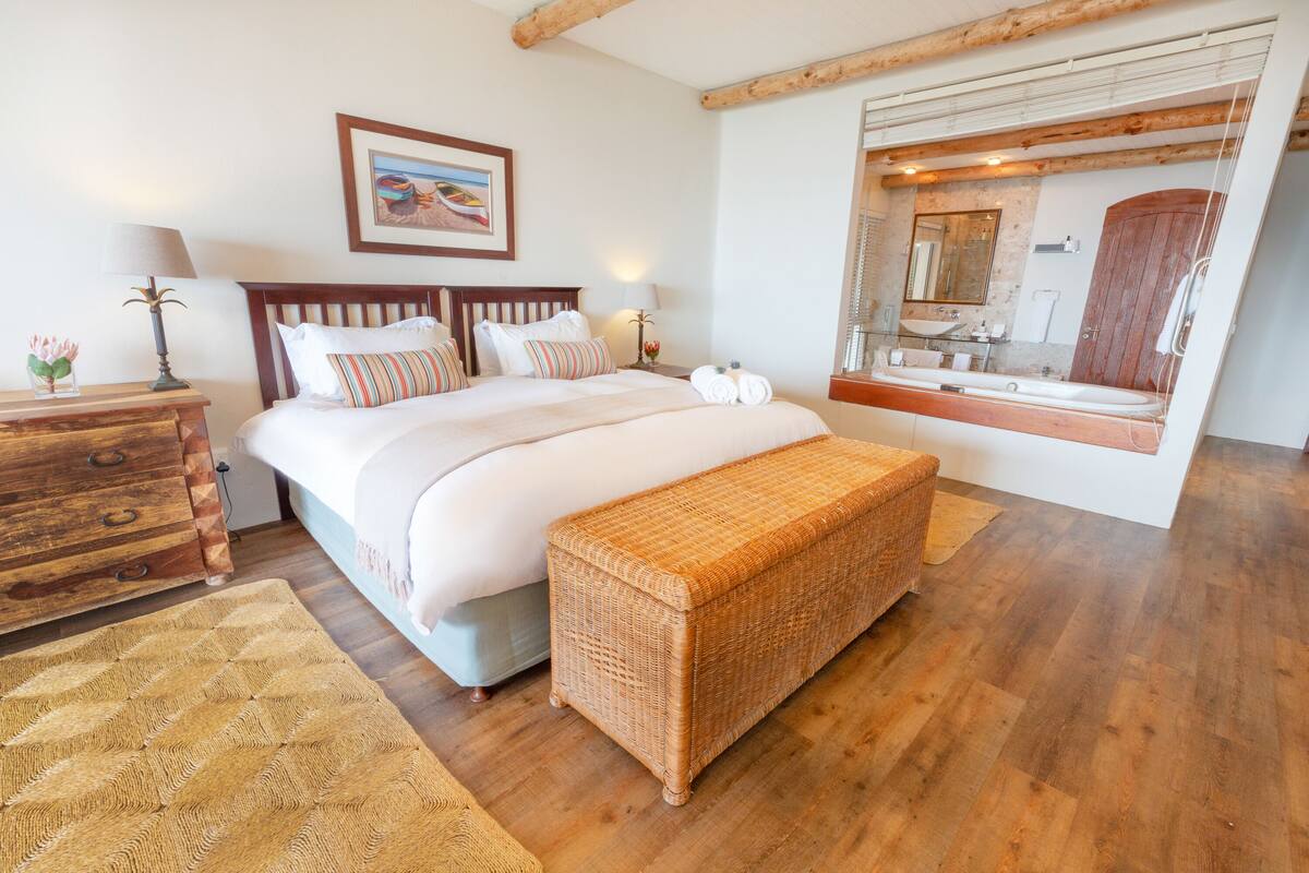 Un dormitorio con una cama grande, cabecera de madera, mesita de noche con lámpara, banco de mimbre y un espejo grande sobre el lavabo.