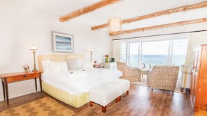 A bedroom with a large bed, wooden beams, a view of the sea, a wicker chair, and a wooden cabinet. - The Sands @ St Francis (Saint Francis Bay)