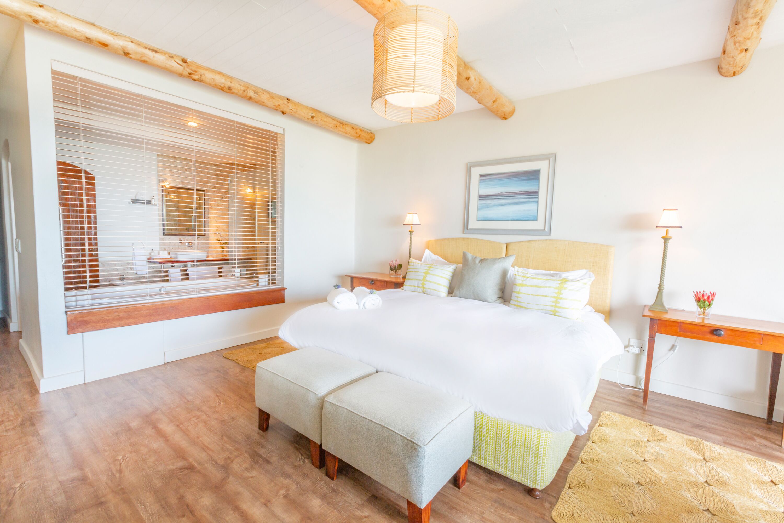 A bedroom with a large bed, wooden beams, a window with blinds, a bedside table with a lamp, and a painting on the wall.