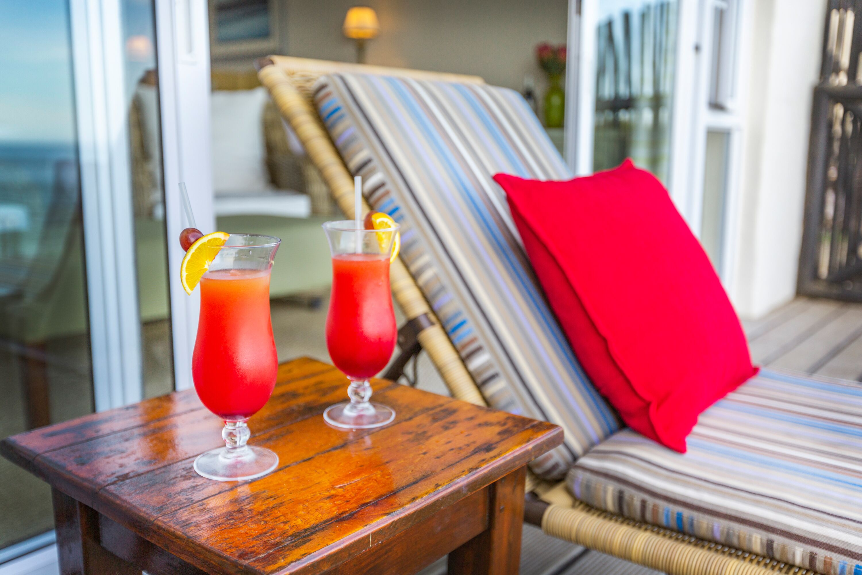Dos bebidas sobre una mesa de madera, con vista a un balcón y una silla a rayas con un cojín rojo.
