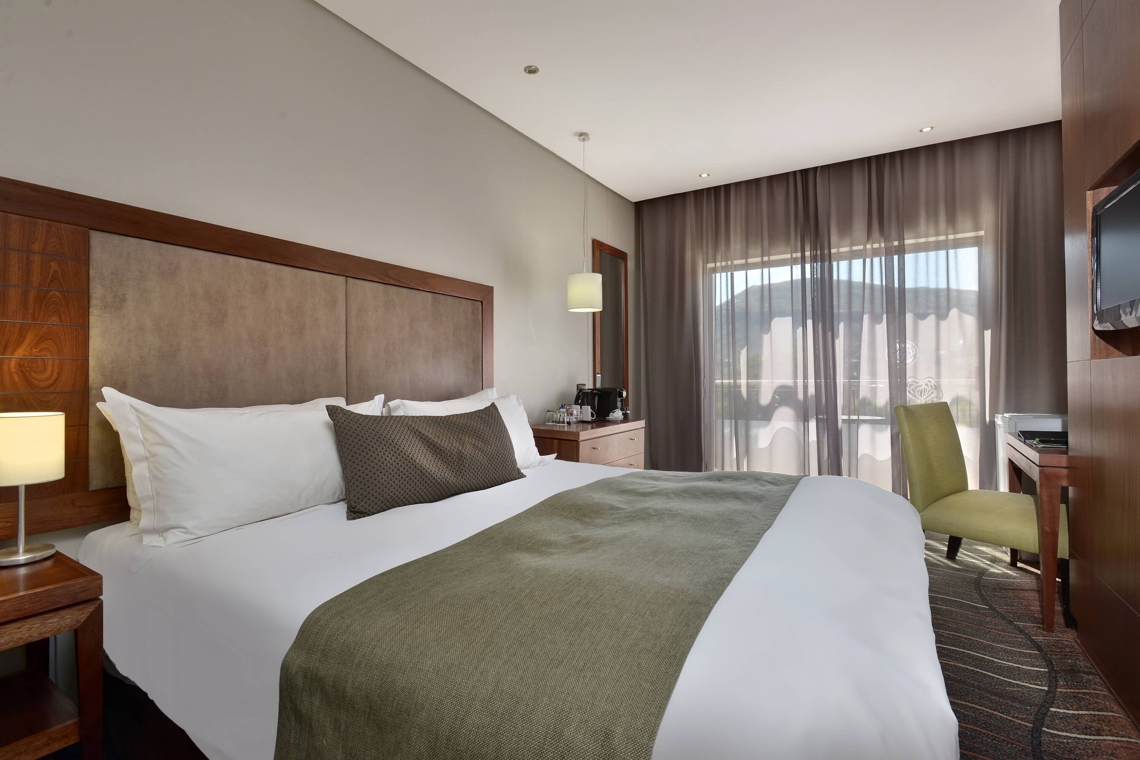 A hotel room with a large bed, a desk with a chair, a TV mounted on the wall, and a window with curtains.