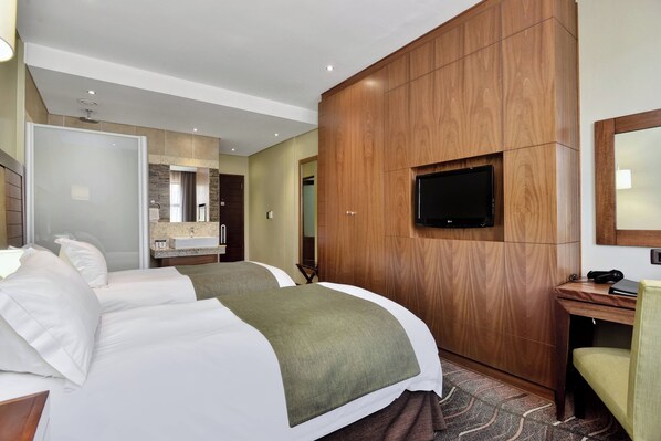 A hotel room with two beds, a wooden wardrobe, a television mounted on the wall, and a small desk with a chair.