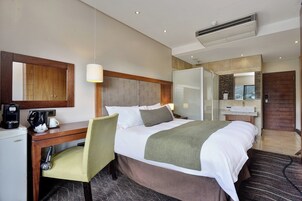 Habitación de hotel con cama, escritorio, silla, cafetera, espejo y zona de ducha.