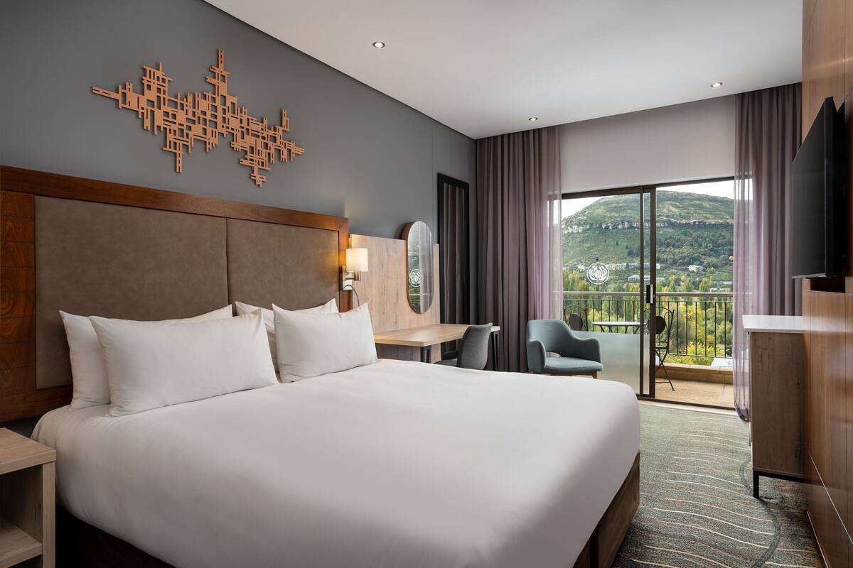 Ein Hotelzimmer mit einem großen Bett, einem Schreibtisch, einem Sessel, einem Fernseher und einem Balkon mit Blick auf die Hügel.