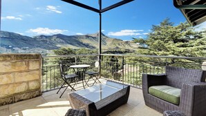 Uma varanda com móveis de vime, uma mesa de vidro e vista para montanhas.