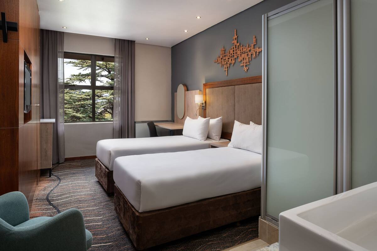 Ein Hotelzimmer mit zwei Betten, einem Schreibtisch, einem Stuhl, einer Badewanne und einem großen Fenster mit Blick auf Bäume.