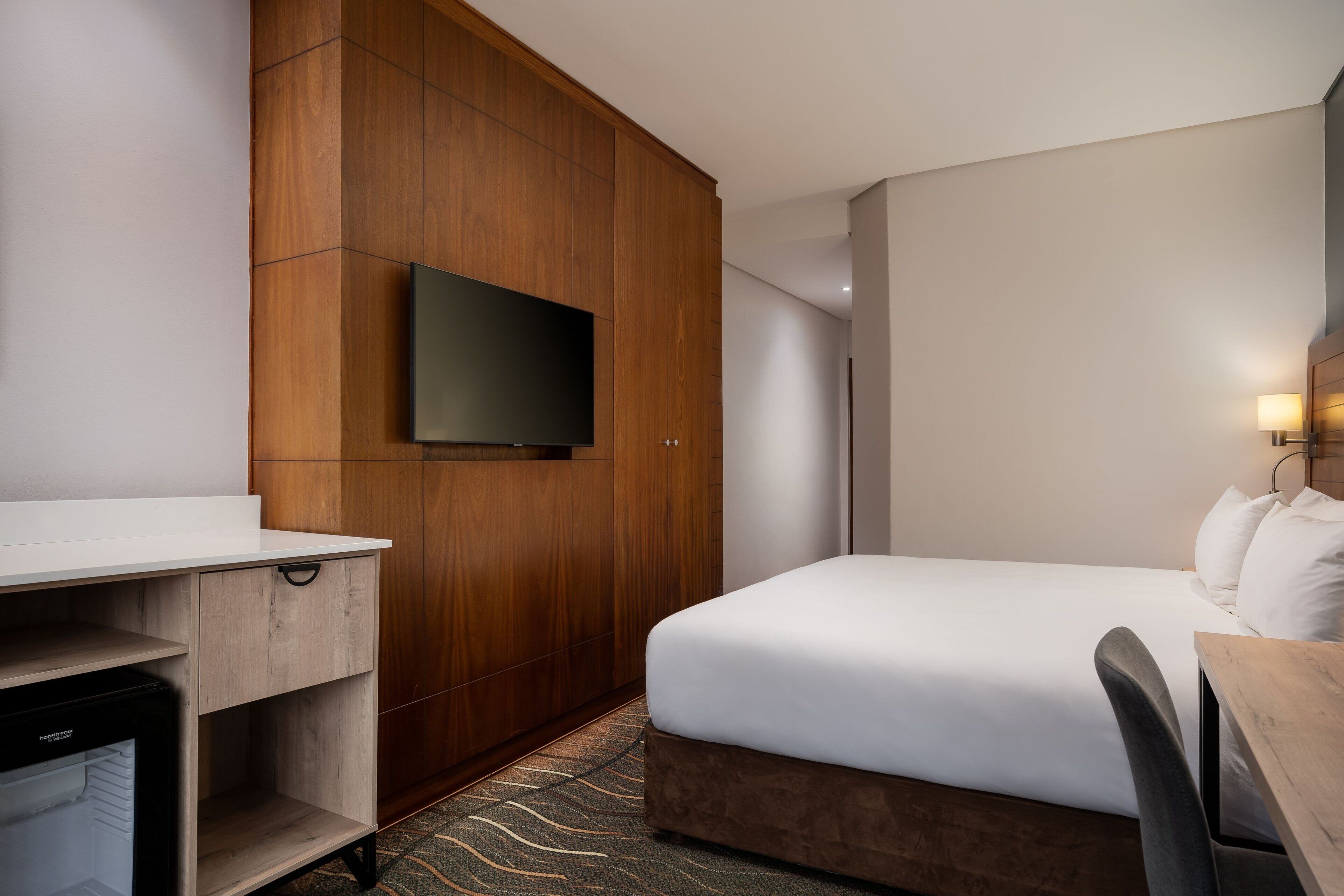 A hotel room with a wooden wardrobe, a flat-screen TV mounted on the wall, a bed with white bedding, and a desk with a chair.