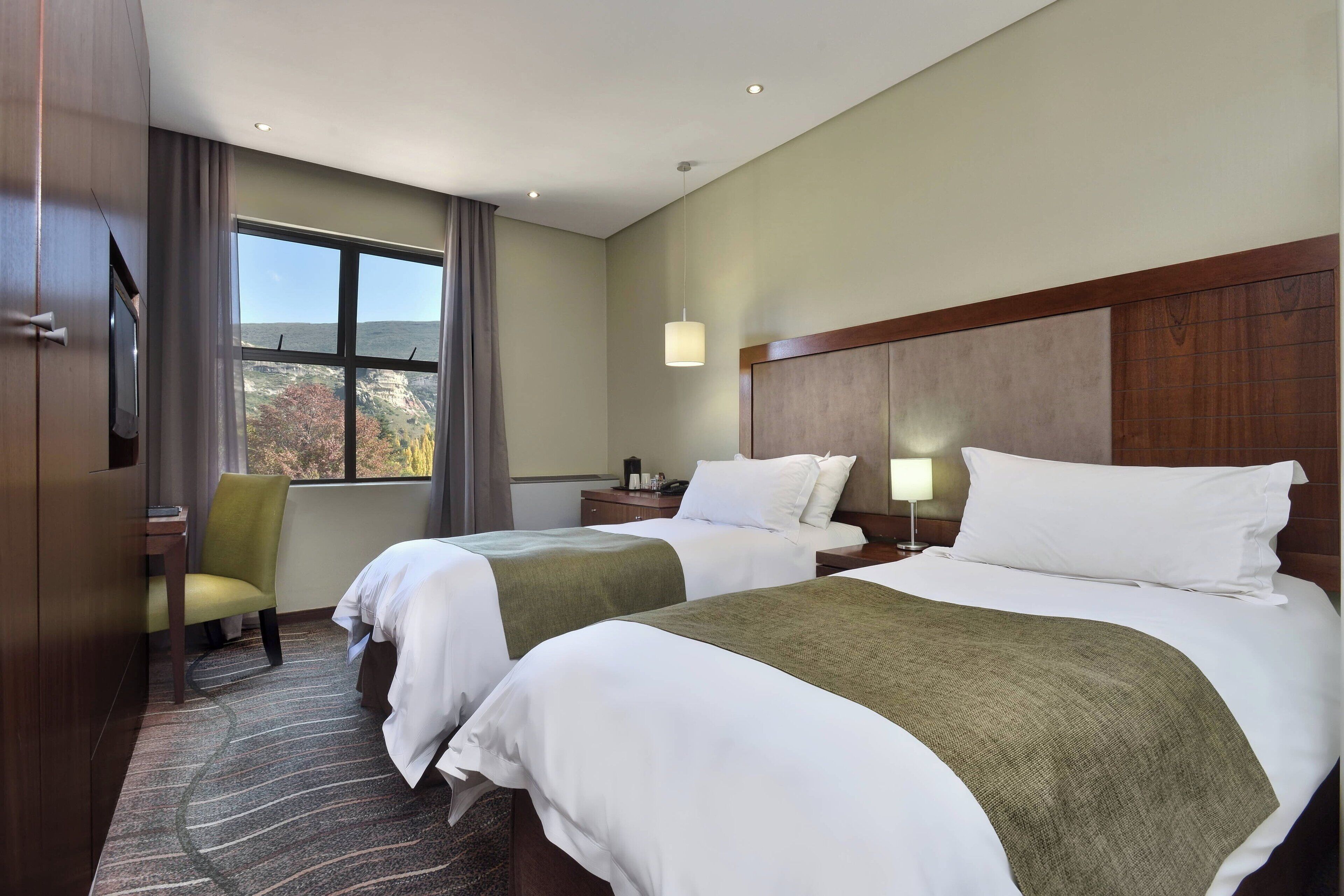Een hotelkamer met twee bedden, een bureau, een stoel, een raam met uitzicht en een lamp.