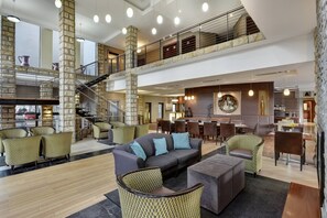 Un hall d’entrée spacieux avec un plafond haut, des piliers en pierre, un escalier et un coin repas comprenant plusieurs tables et chaises.