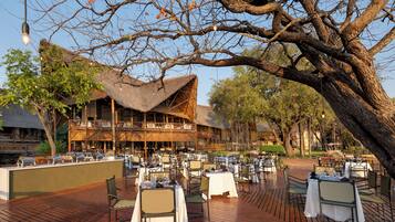 An outdoor dining area with tables and chairs, a thatched-roof building, and a large tree in the background.