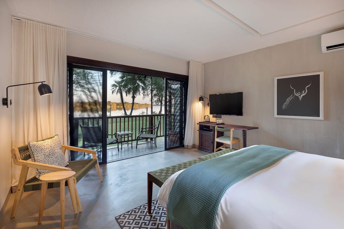 Quarto de hotel com cama, secretária com televisão, cadeira, varanda com vista para palmeiras e janela com cortinas.