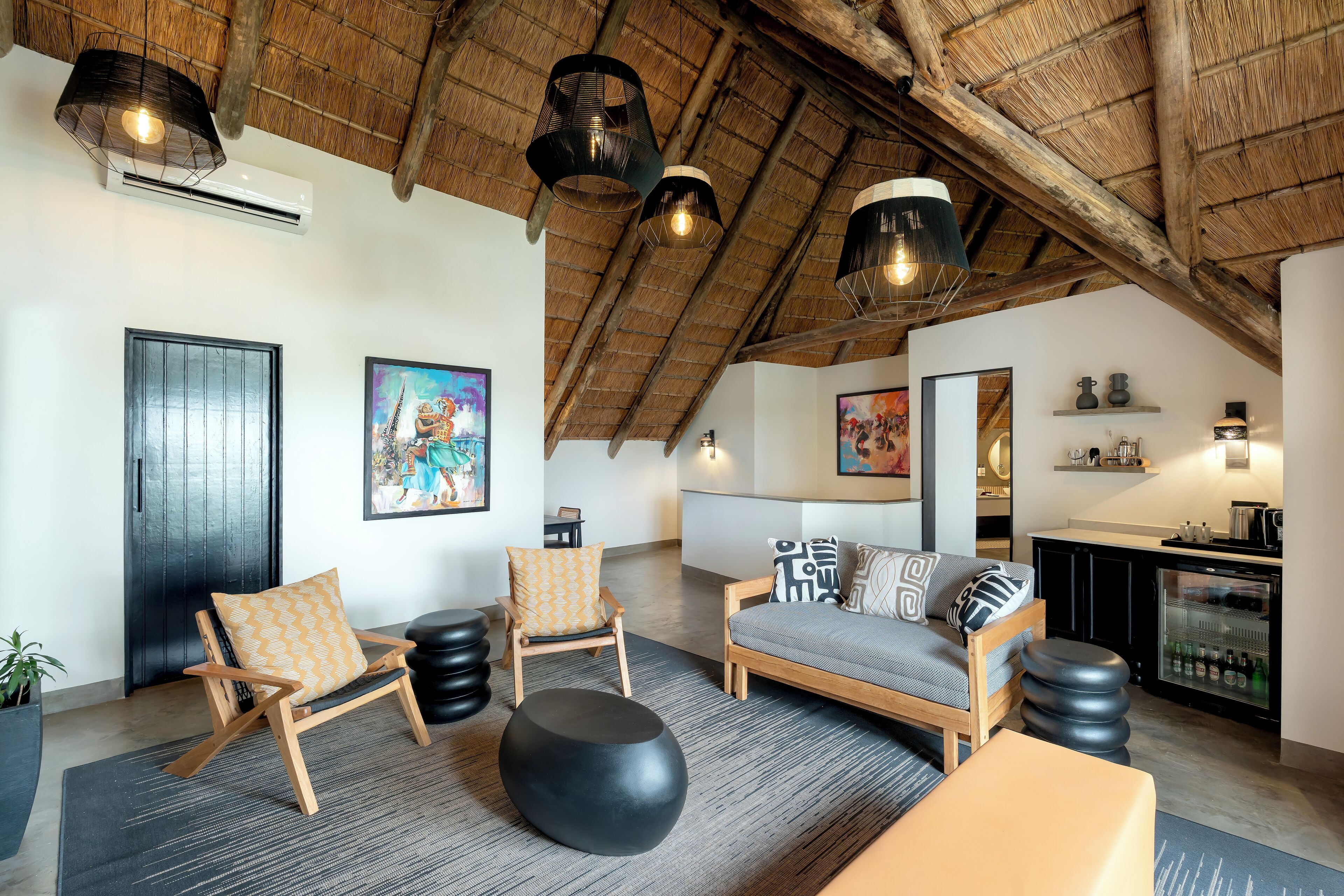 A spacious loft with a high ceiling, exposed wooden beams, a cozy seating area with a sofa and chairs, a small kitchenette, and artwork on the walls.