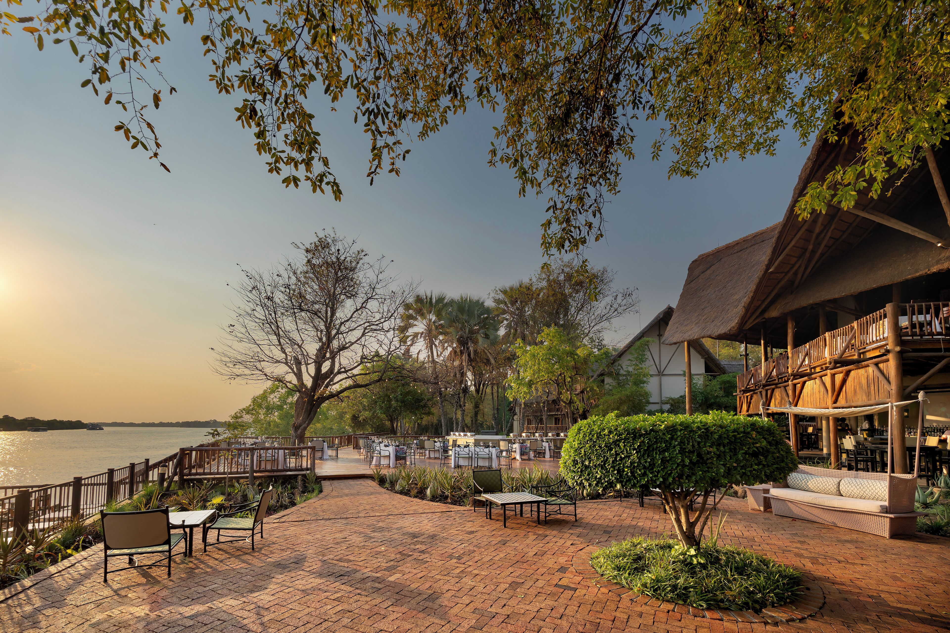 A riverside patio with seating, a wooden deck, and a building with a thatched roof.
