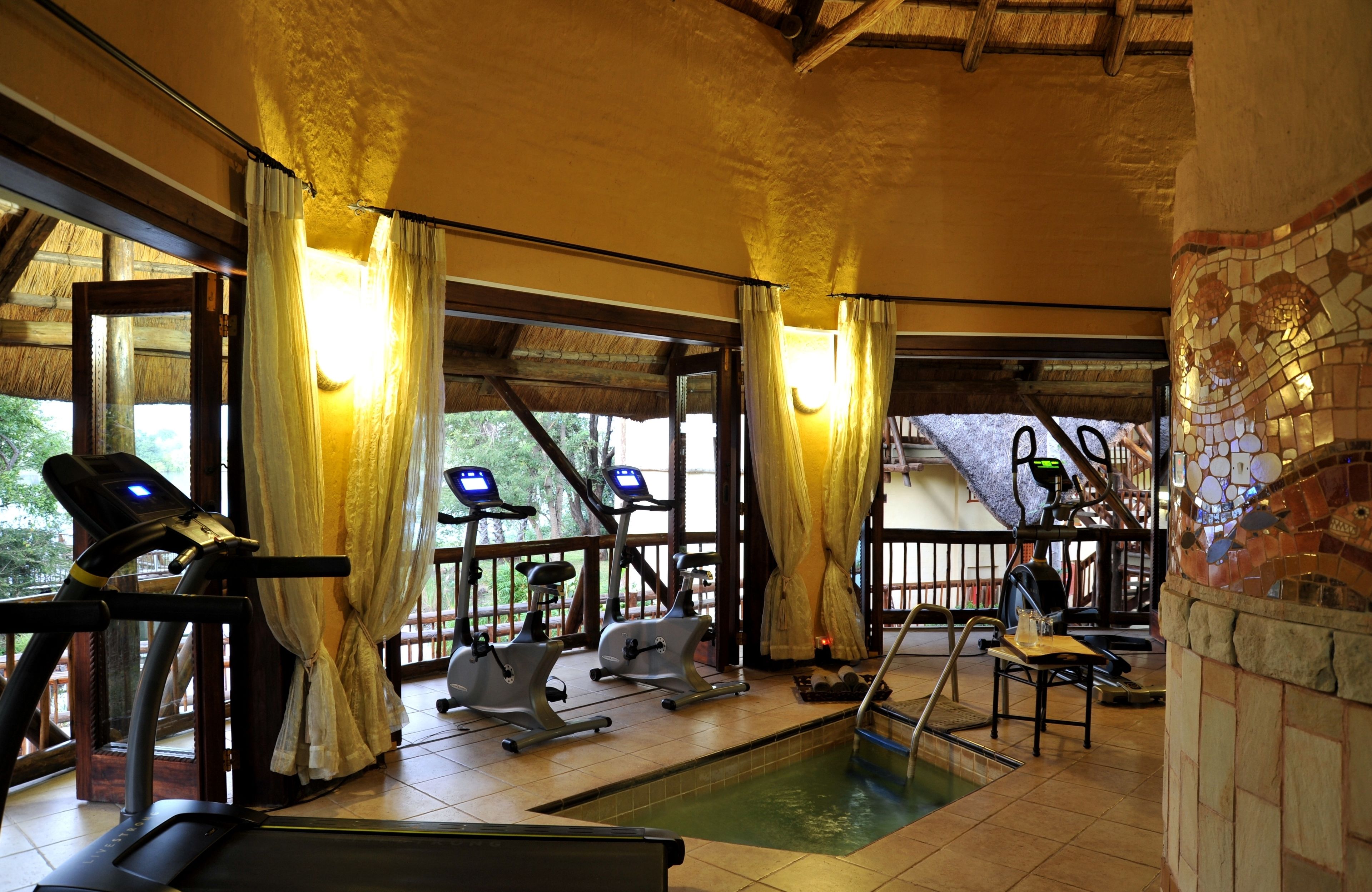 Un gimnasio con equipamiento de ejercicio, piscina y ventanas grandes con cortinas.