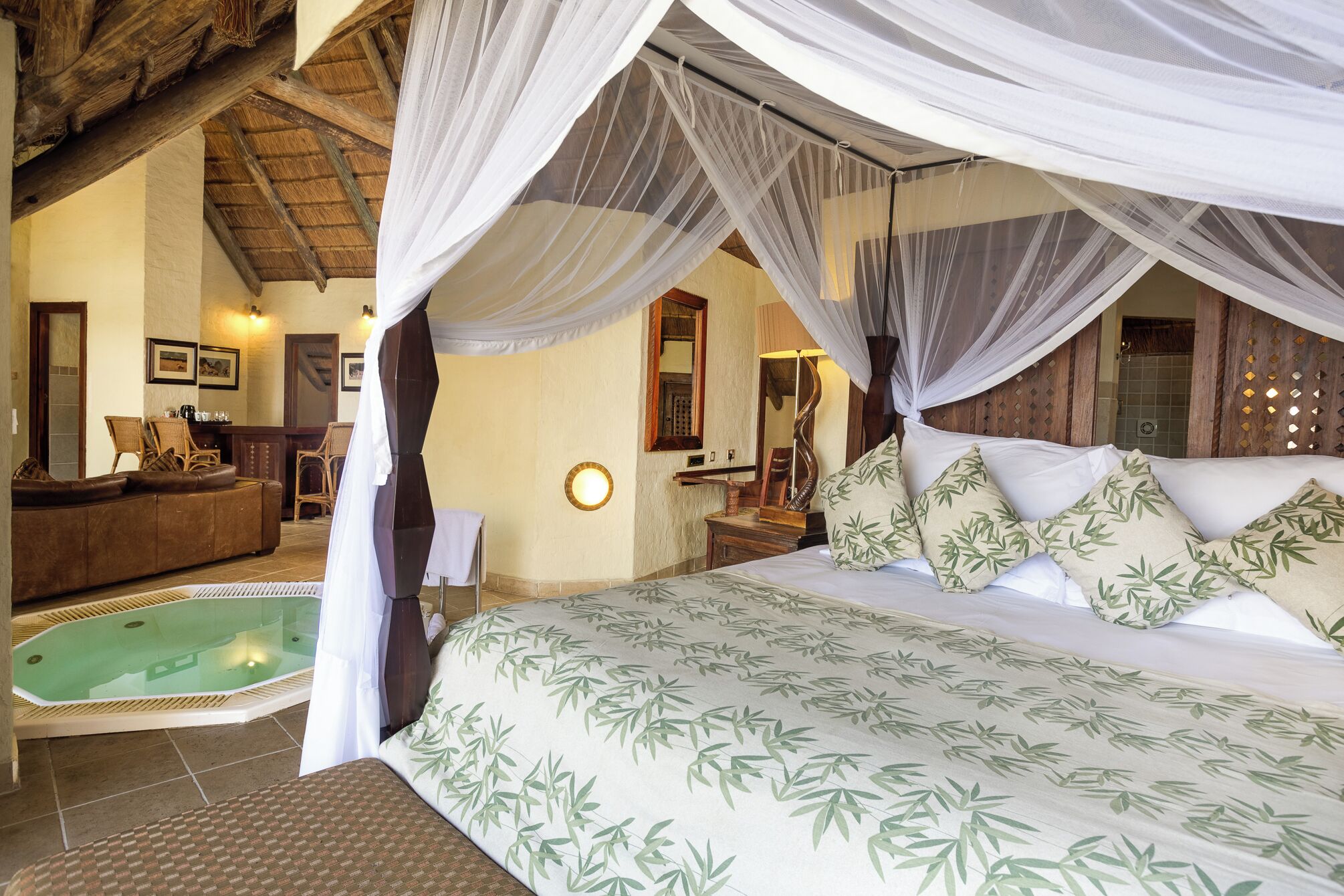 A bedroom with a canopy bed, a sitting area with a sofa and chairs, a small pool, and a bathroom with a mirror and sink.
