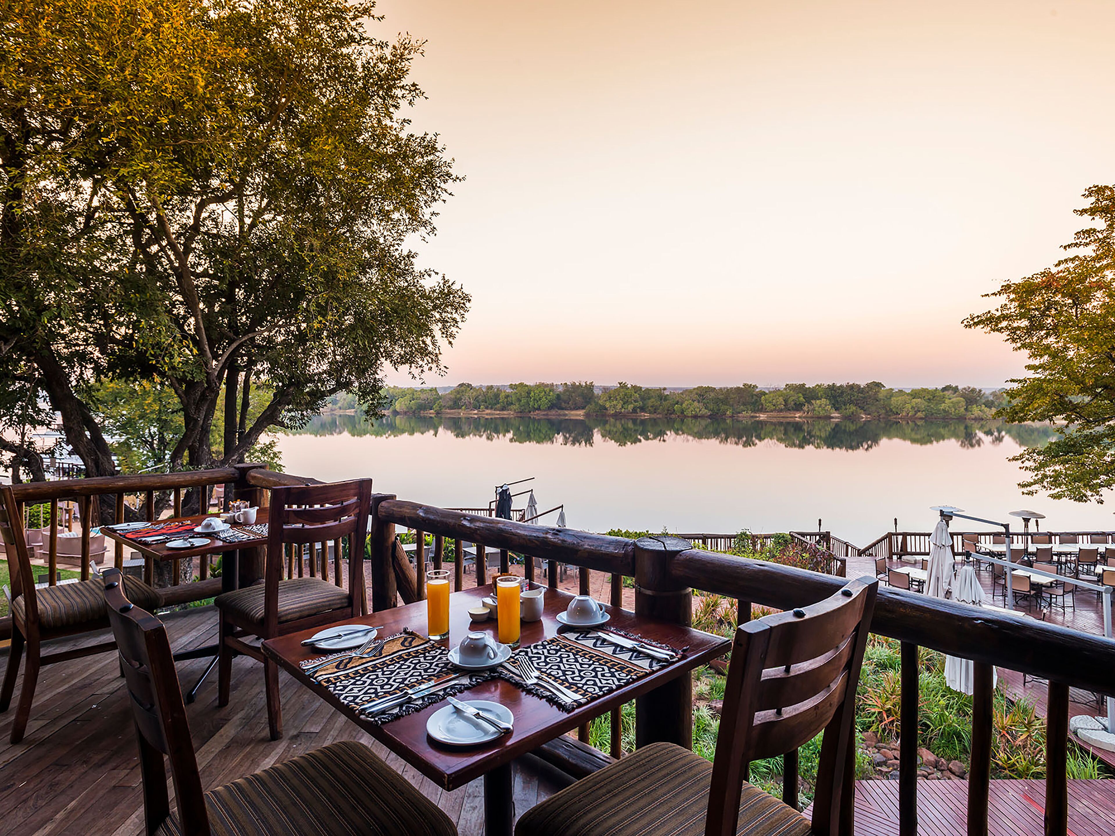A dining area with a table set for two, overlooking a calm lake with trees in the background.