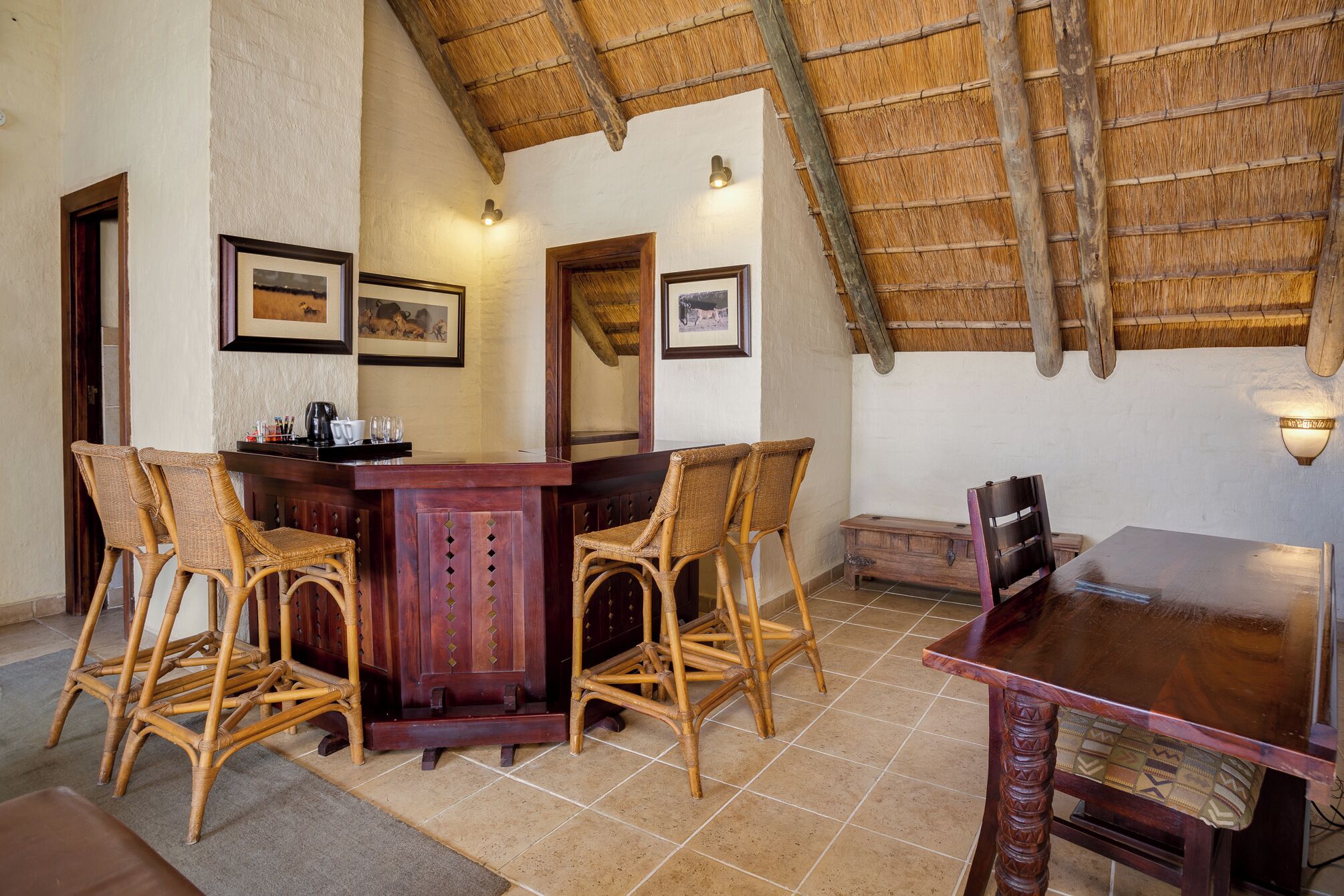 A dining area with a wooden table and four wicker chairs, a bar area with a counter and bar stools, and a high thatched ceiling.