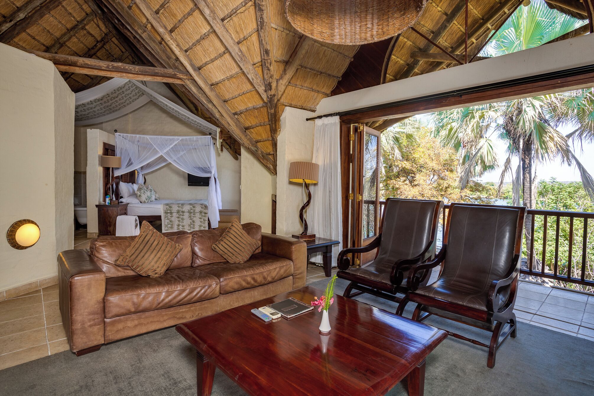 A room with a leather sofa, a wooden coffee table, and two leather chairs. There is a bed with a canopy and a nightstand with a lamp. The room has a large window with a view of palm trees.