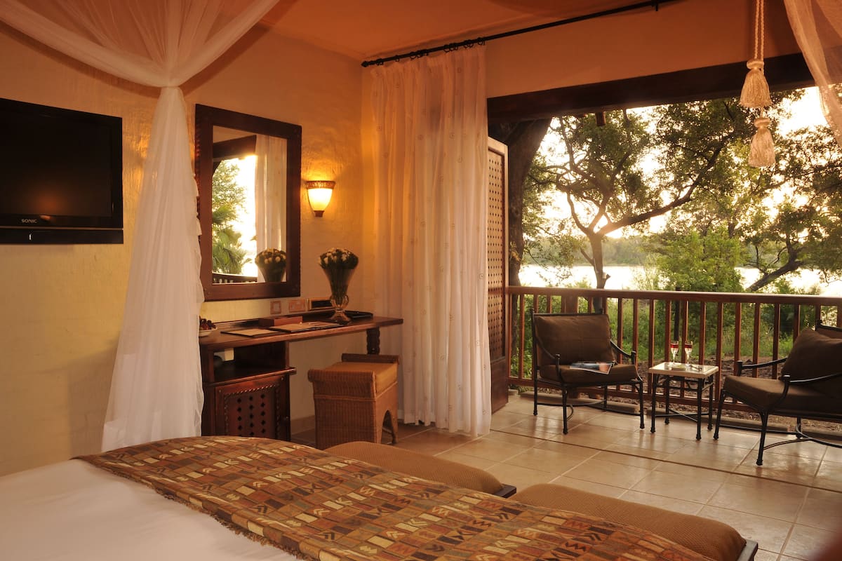 Quarto com cama, escrivaninha e cadeira, varanda com vista para árvores e um corpo de água.
