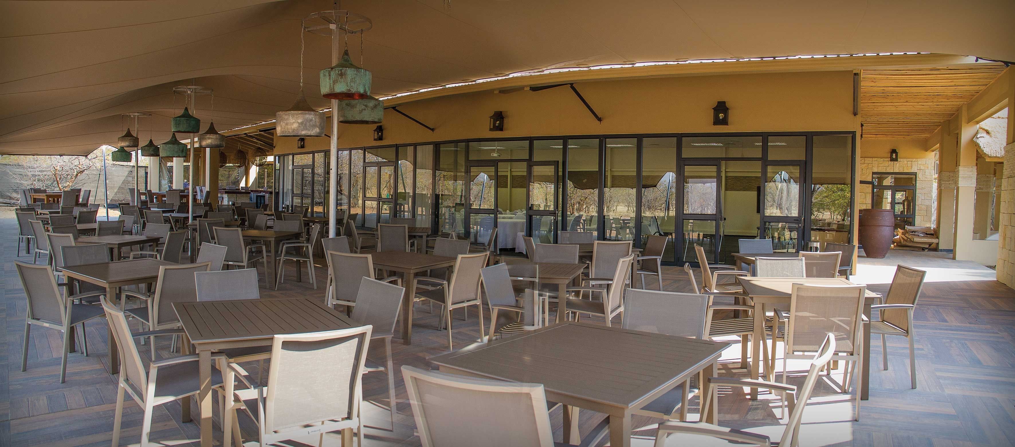 An outdoor cafe with multiple tables and chairs, large windows, and hanging light fixtures.