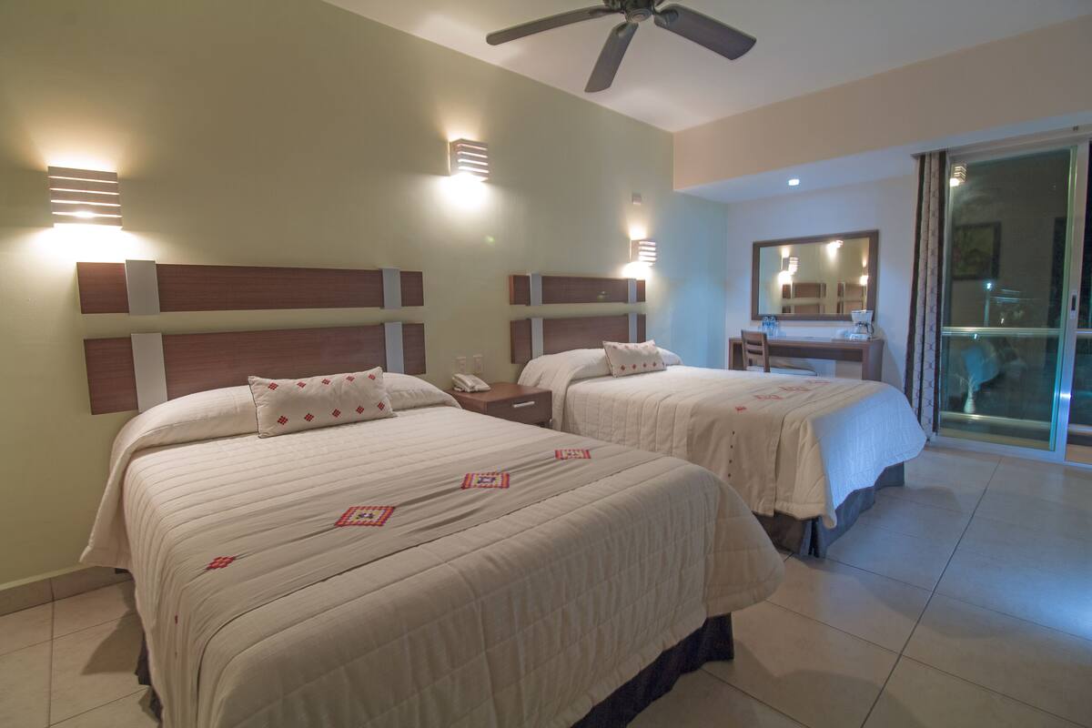 Habitación de hotel con dos camas, un escritorio, una silla, un ventilador de techo y un ventanal corredero.