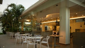 Un área de comedor al aire libre con sillas y mesas blancas, palmeras y un edificio moderno con paredes de vidrio.