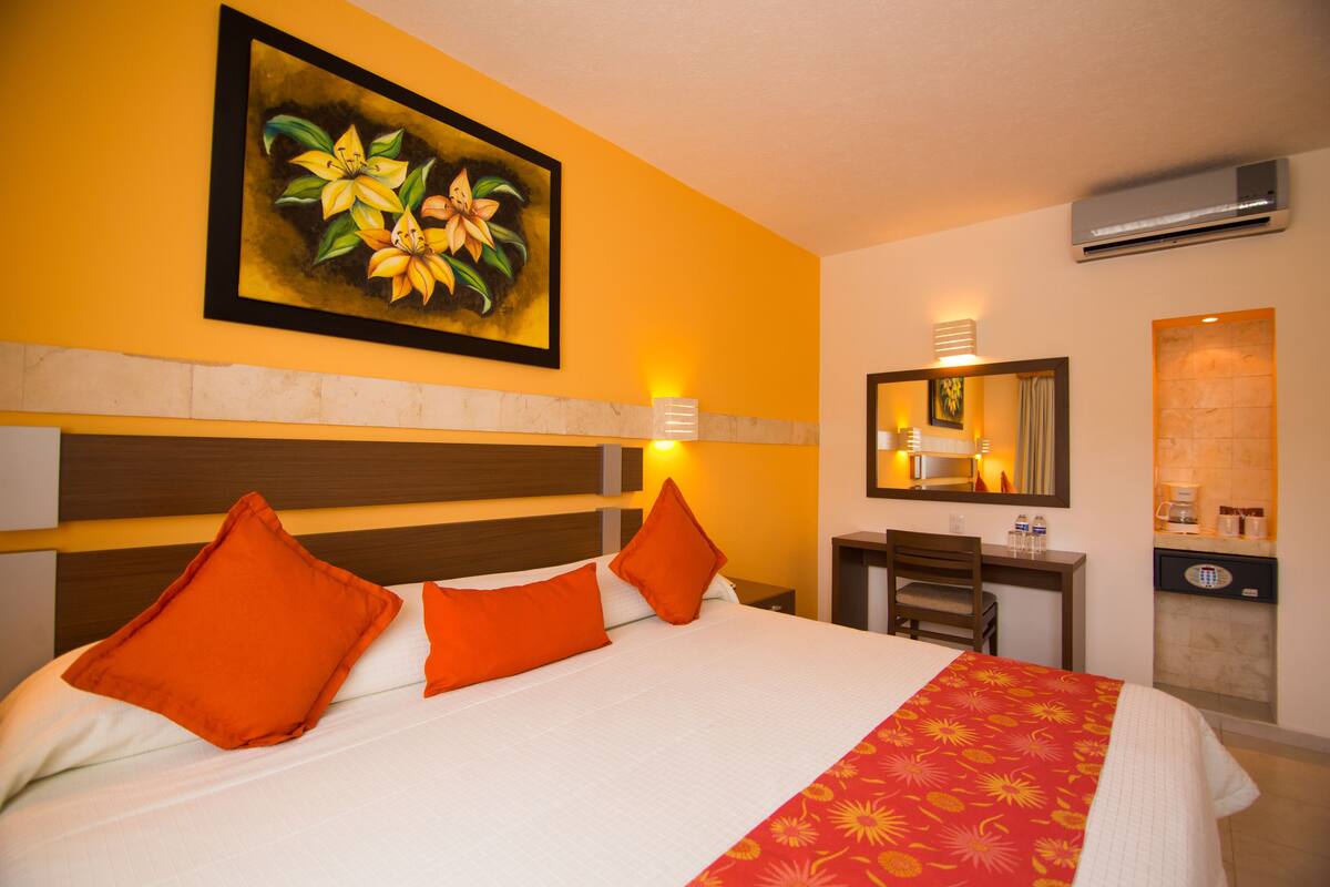 Habitación de hotel con cama, almohadas naranjas, un cuadro de flores, escritorio con silla, espejo y aire acondicionado.