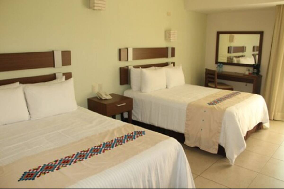 Habitación de hotel con dos camas, una mesita de noche con lámpara, un espejo y un pequeño escritorio con silla.