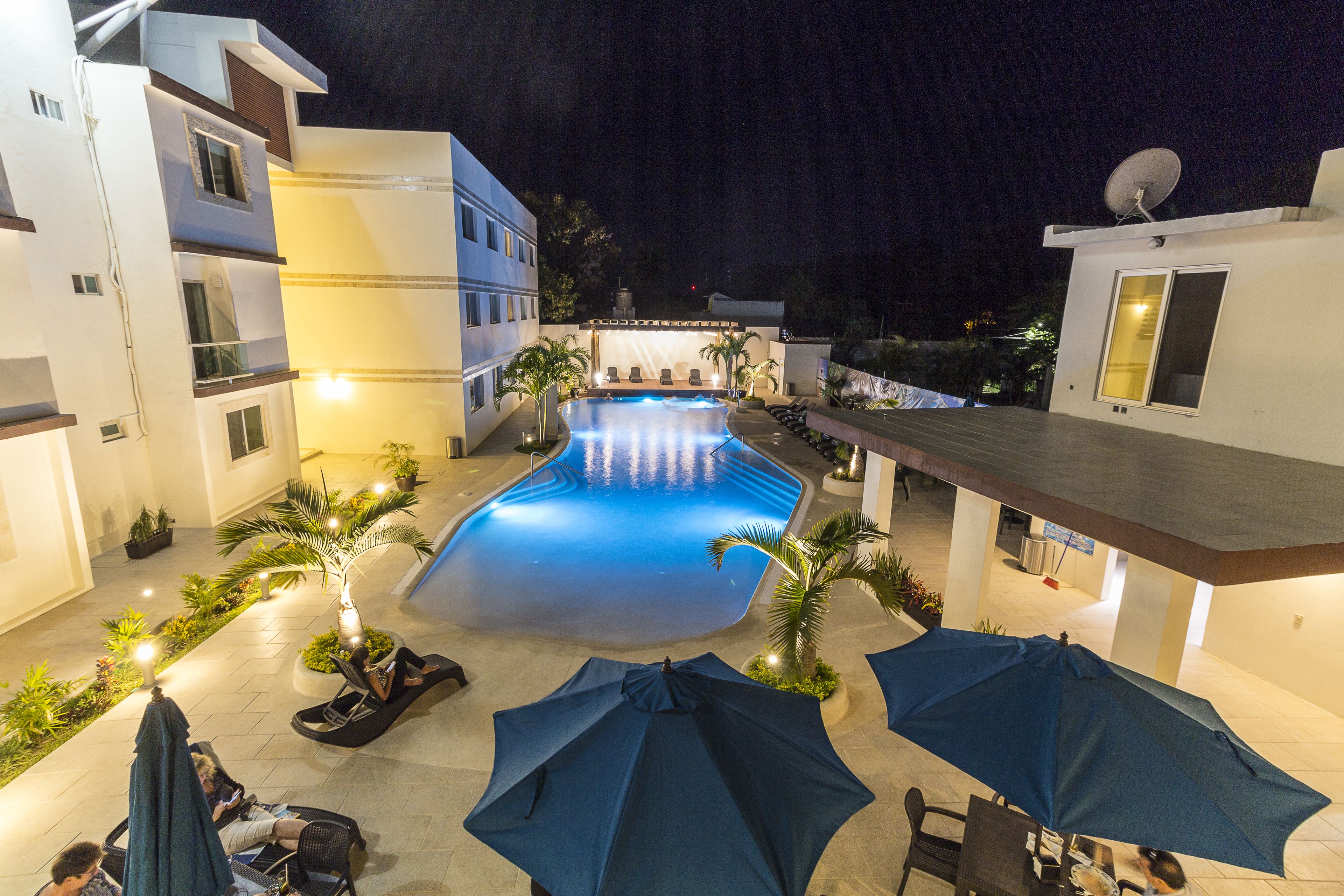 A swimming pool area with lounge chairs and umbrellas, surrounded by buildings with balconies and a satellite dish.