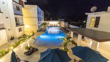 A swimming pool area with lounge chairs and umbrellas, surrounded by buildings with balconies and a satellite dish.
