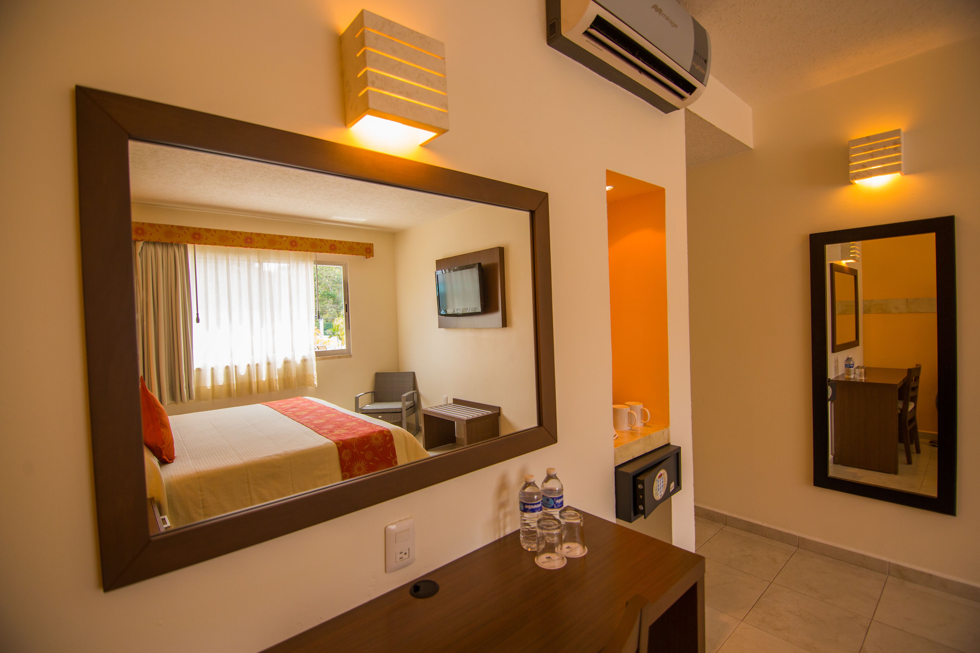 Una habitación de hotel con un espejo grande, una cama con almohada roja, un televisor, un escritorio con botellas de agua y un equipo de aire acondicionado.