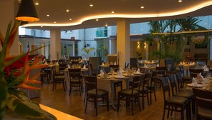 Un restaurante espacioso con varias mesas y sillas, ventanas amplias y plantas de interior.