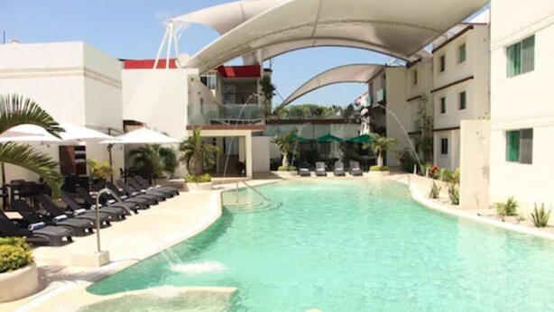 Una piscina con sillas de descanso y sombrillas, rodeada de edificios con balcones.