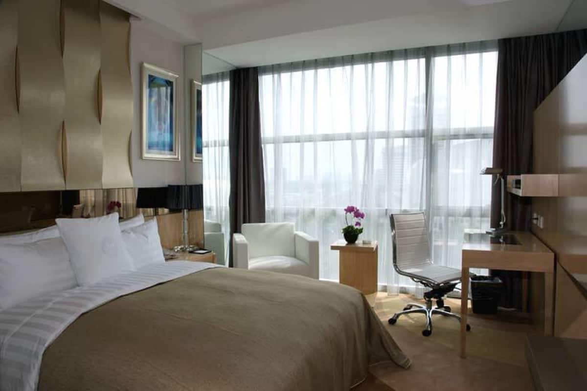 A hotel room with a large bed, a desk, a chair, and a view of the city through sheer curtains.