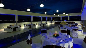 A spacious outdoor dining area with multiple tables set for guests, large windows, and a view of the city at night. - Kapila Business Hotel (Pune)