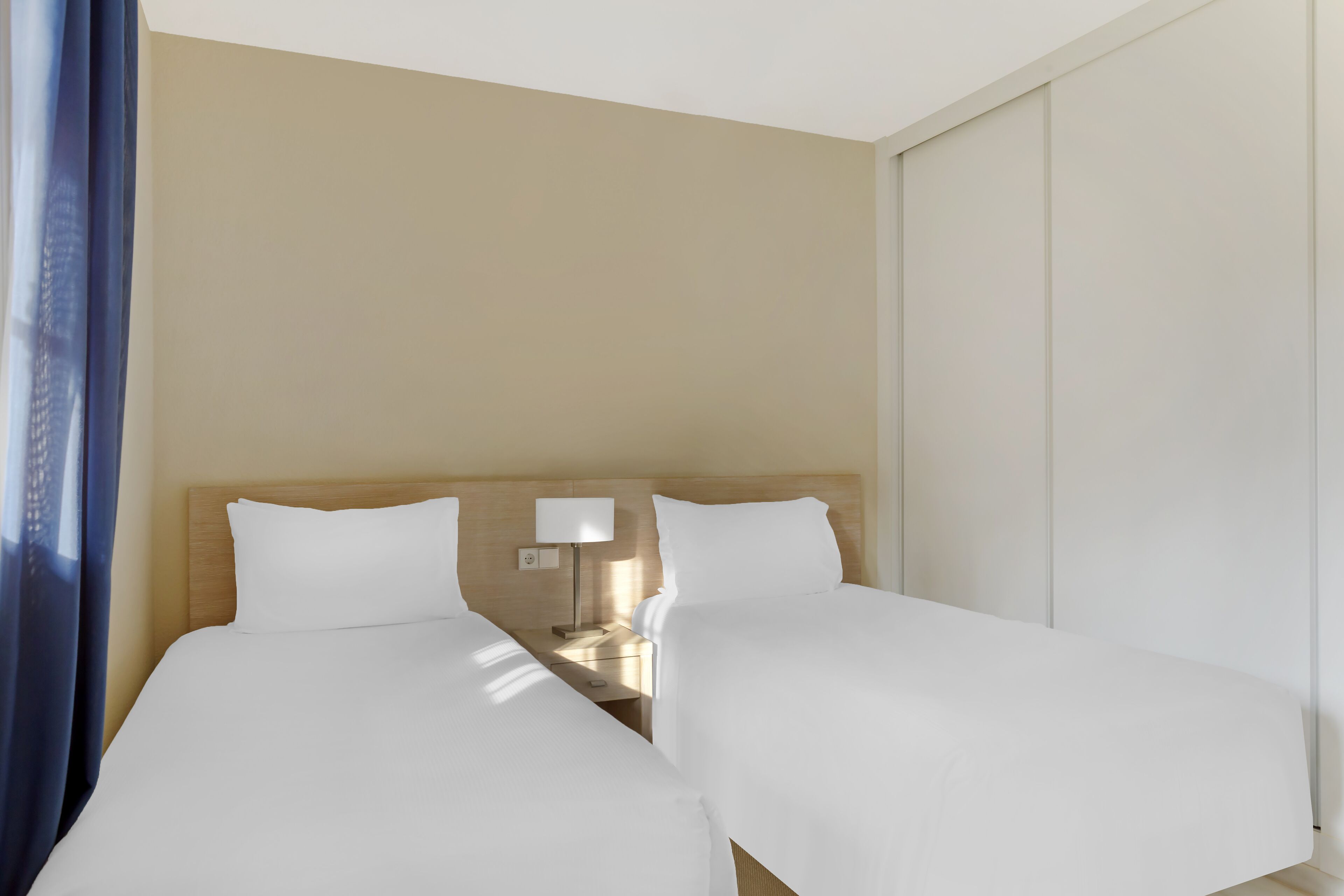 A hotel room with two beds, a nightstand with a lamp, and a view of the city through the window.
