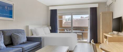 A hotel room with a bed, a sofa, a dining table, a TV, and a view of the outside.